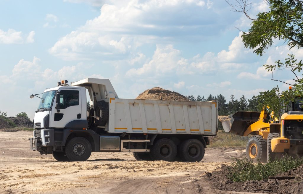 Dump truck with a bunch of land in the back and excavator at the construction site outside the city. Road works on an intercity highway on a cloudy day. Selective focus Dump truck with a bunch of land in the back and excavator at the construction site outside the city. Road works on an intercity highway on a cloudy day. Selective focus